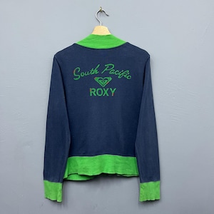 May include: A navy blue long-sleeve shirt with a bright green mock neck collar, cuffs, and hem. The back features the green text South Pacific and ROXY with a heart-shaped logo in the center. The shirt is displayed on a wooden hanger.