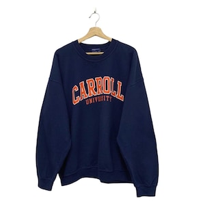 以下が含まれることがあります： オレンジ色の文字で「Carroll University」と書かれたネイビーブルーのスウェットシャツ。白い背景に木製のハンガーに吊るされています。クルーネックと長袖です。