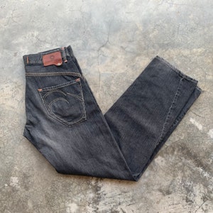 May include: Dark grey denim jeans with a straight leg cut. Features a brown leather patch on the back waistband and a unique white stitched design on the back pocket. Orange stitching accents the seams.