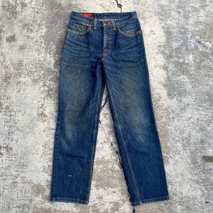 Pode incluir: Um par de jeans vintage de ganga azul com braguilha de bot&otilde;es e design cl&aacute;ssico de cinco bolsos. Os jeans mostram sinais de desgaste, com &aacute;reas desbotadas e um visual desgastado. Corte reto e uma etiqueta vermelha no bolso de tr&aacute;s.