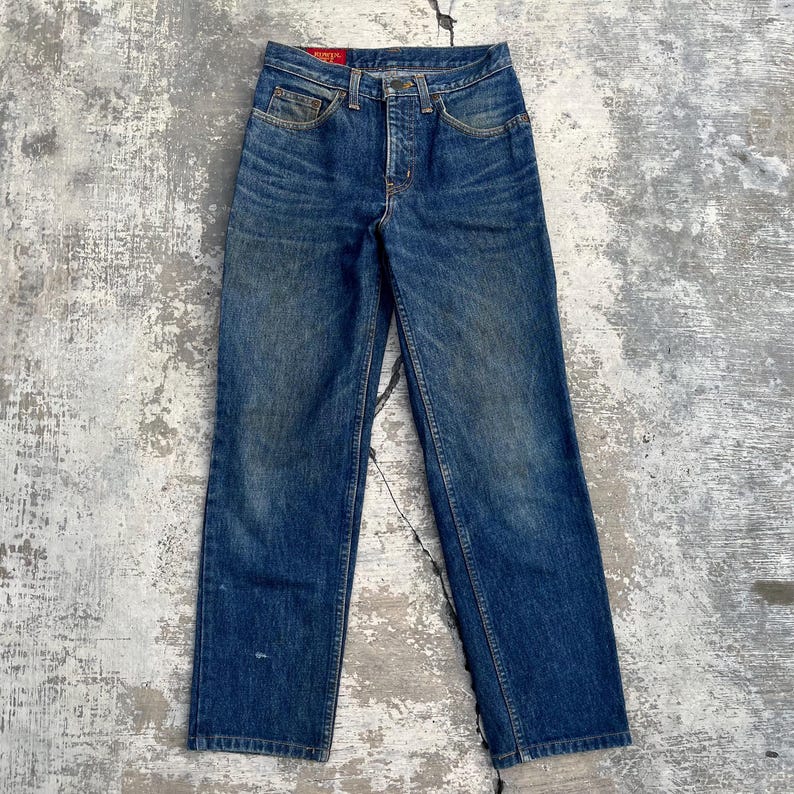 Pode incluir: Um par de jeans vintage de ganga azul com braguilha de bot&otilde;es e design cl&aacute;ssico de cinco bolsos. Os jeans mostram sinais de desgaste, com &aacute;reas desbotadas e um visual desgastado. Corte reto e uma etiqueta vermelha no bolso de tr&aacute;s.