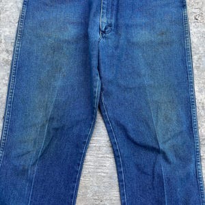 May include: Close-up of blue denim jeans. The jeans have a classic five-pocket design, a button and zipper closure, and a faded appearance. The denim has some discoloration and wear.