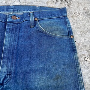 May include: Close-up of blue denim jeans. The jeans have a button closure, belt loops, and front pockets. The denim has a faded appearance with some wear and tear, and the metal rivets and button are visible.