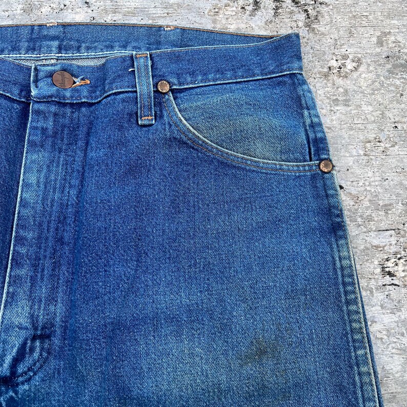 May include: Close-up of blue denim jeans. The jeans have a button closure, belt loops, and front pockets. The denim has a faded appearance with some wear and tear, and the metal rivets and button are visible.