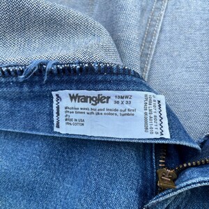 May include: Close-up of a pair of blue denim Wrangler jeans. The label reads "Wrangler 13MWZ 36 X 33" and includes washing instructions and "MADE IN USA" text. The jeans are made of 100% cotton.