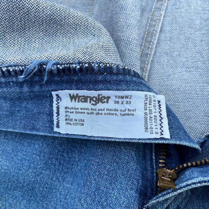 May include: Close-up of a pair of blue denim Wrangler jeans. The label reads "Wrangler 13MWZ 36 X 33" and includes washing instructions and "MADE IN USA" text. The jeans are made of 100% cotton.