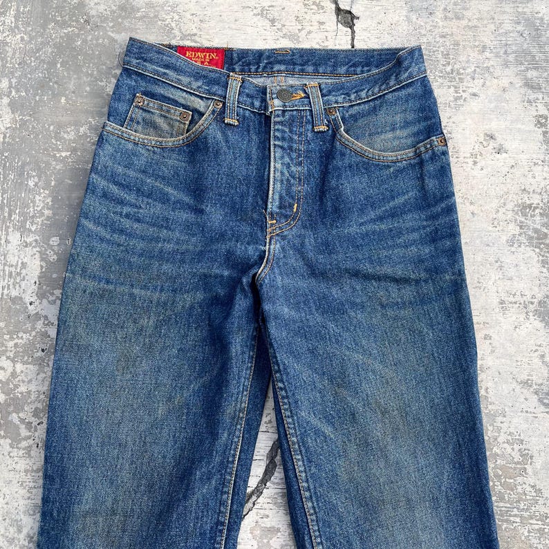 Pode incluir: Cal&ccedil;as de ganga azuis com lavagem desbotada. As cal&ccedil;as de ganga t&ecirc;m um design cl&aacute;ssico de cinco bolsos, braguilha de bot&otilde;es e uma etiqueta vermelha Edwin. As cal&ccedil;as de ganga parecem vintage, com algum desgaste vis&iacute;vel no tecido.