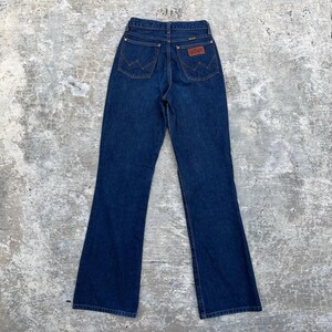 K&ouml;nnte beinhalten: Dunkelblaue Wrangler Jeans aus Denim mit hoher Taille und ausgestelltem Bein. Die Jeans haben ein klassisches F&uuml;nf-Taschen-Design mit dem Wrangler-Logo auf den Ges&auml;&szlig;taschen und einem braunen Lederaufn&auml;her.