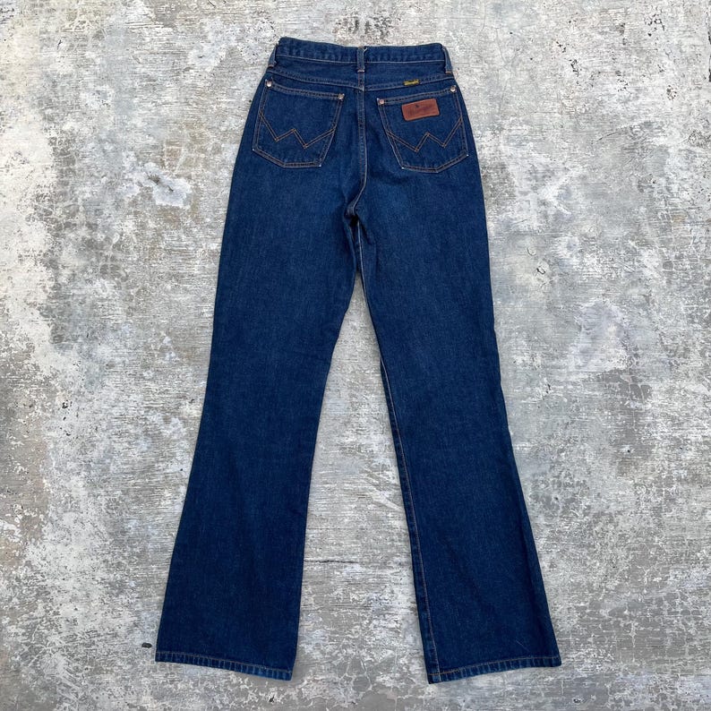 K&ouml;nnte beinhalten: Dunkelblaue Wrangler Jeans aus Denim mit hoher Taille und ausgestelltem Bein. Die Jeans haben ein klassisches F&uuml;nf-Taschen-Design mit dem Wrangler-Logo auf den Ges&auml;&szlig;taschen und einem braunen Lederaufn&auml;her.