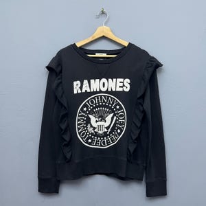 May include: Black sweatshirt with the word "RAMONES" in white block letters. The sweatshirt features a circular graphic with the names "JOHNNY", "JOEY", "DEEDEE", and "TOMMY" around a central emblem. Ruffled trim accents the shoulders.