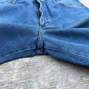 May include: Close-up of blue denim jeans. The jeans have a zipper fly and classic five-pocket design. The fabric shows subtle fading and wear, suggesting a vintage or well-loved item. The stitching is visible along the seams.