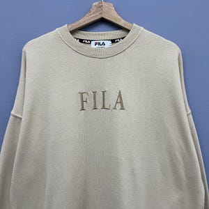 May include: A beige crewneck sweatshirt featuring the word FILA embroidered in a matching thread color on the chest. The interior collar displays a black branded tape with the FILA logo repeated in white and red text, along with a white brand label.