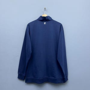 Puede incluir: Sudadera azul marino de manga larga con cuello alzado y un peque&ntilde;o logotipo blanco en la parte superior de la espalda. La sudadera est&aacute; colgada de una percha met&aacute;lica sobre un fondo azul claro. La sudadera tiene una cremallera que sube hasta el cuello alzado.