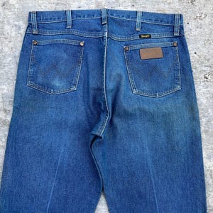 May include: Close-up of the back of blue denim jeans. The jeans have two back pockets with the Wrangler logo stitched on them. The jeans have a brown leather patch with the Wrangler logo on the back right pocket.
