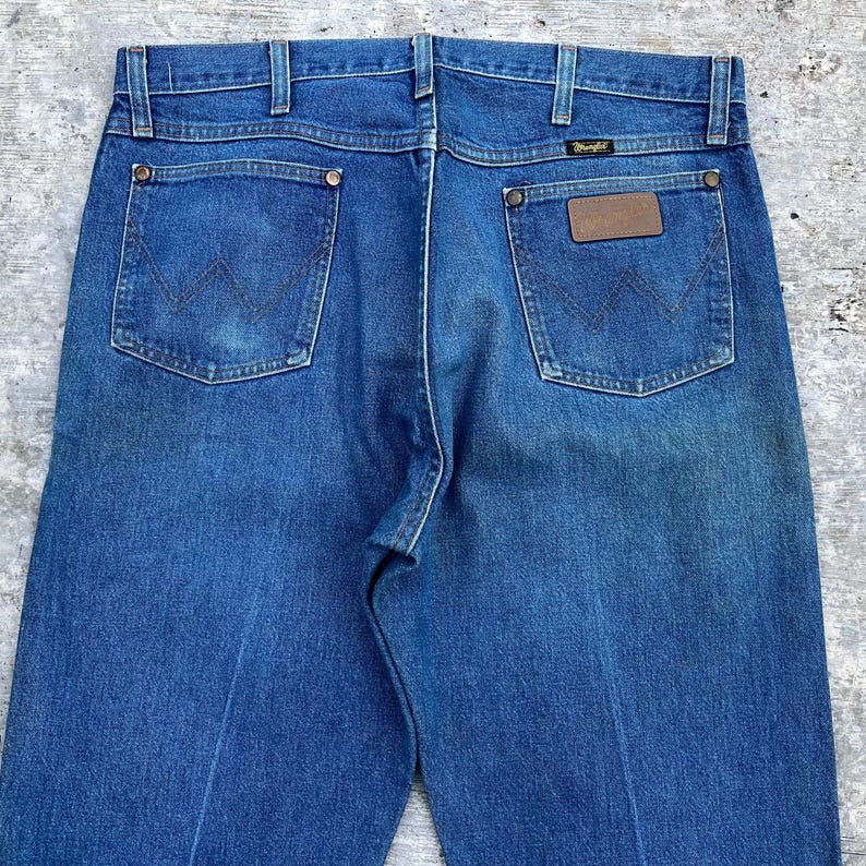 May include: Close-up of the back of blue denim jeans. The jeans have two back pockets with the Wrangler logo stitched on them. The jeans have a brown leather patch with the Wrangler logo on the back right pocket.