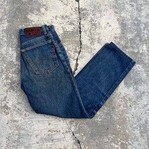 Pode incluir: Cal&ccedil;as de ganga azuis com um design cl&aacute;ssico de cinco bolsos. As cal&ccedil;as de ganga apresentam um remendo de couro castanho com a marca "EDWIN" e uma etiqueta vermelha. O bolso traseiro tem uma costura distinta em forma de "W".
