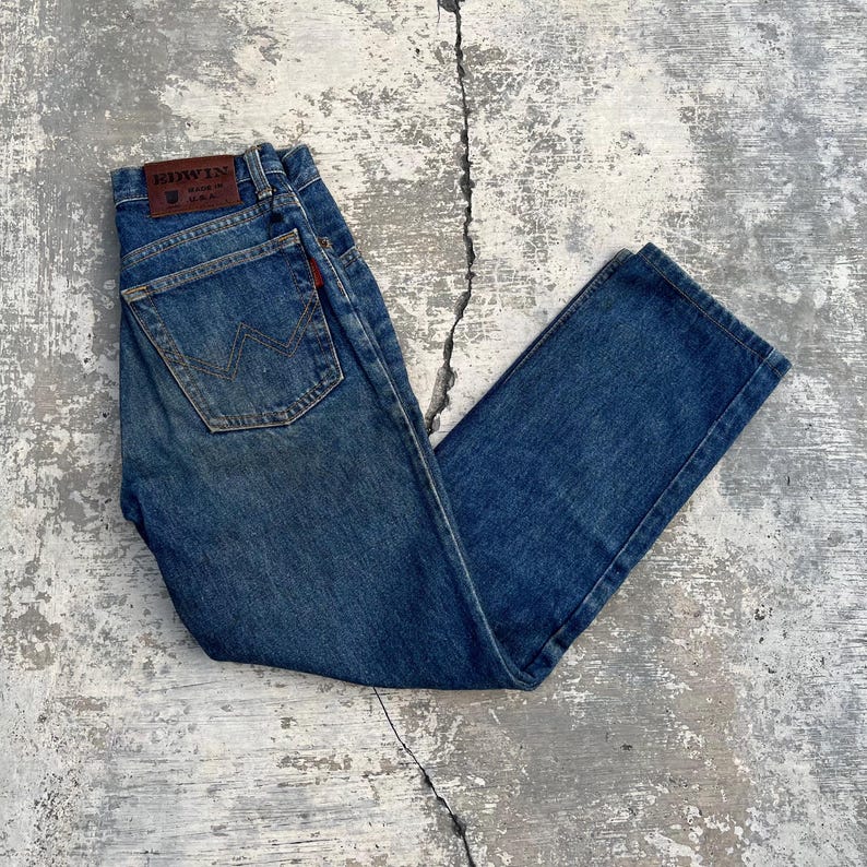 Pode incluir: Cal&ccedil;as de ganga azuis com um design cl&aacute;ssico de cinco bolsos. As cal&ccedil;as de ganga apresentam um remendo de couro castanho com a marca "EDWIN" e uma etiqueta vermelha. O bolso traseiro tem uma costura distinta em forma de "W".