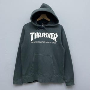 以下が含まれることがあります： ドローコード付きフードとカンガルーポケットが付いた濃いグレーのパーカー。胸には白い文字で「THRASHER SKATEBOARD MAGAZINE」とプリントされています。パーカーは柔らかく快適な素材で作られています。
