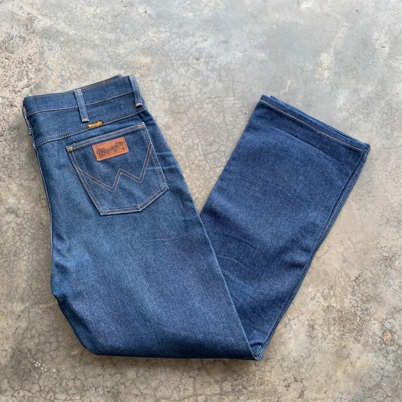 K&ouml;nnte beinhalten: Dunkelblaue Jeans aus Denim mit klassischem F&uuml;nf-Taschen-Design. Die Jeans haben einen braunen Lederaufn&auml;her mit dem Wrangler-Logo auf der Ges&auml;&szlig;tasche. Die Jeans sind gefaltet und flach hingelegt, was den geraden Bein-Schnitt zeigt.
