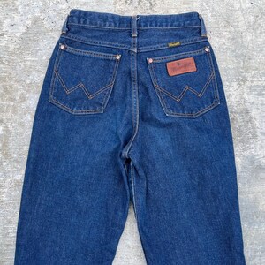 K&ouml;nnte beinhalten: Dunkelblaue Jeans aus Denim mit dem Wrangler-Logo auf der Ges&auml;&szlig;tasche. Die Jeans haben eine hohe Taille, ein klassisches F&uuml;nf-Taschen-Design und kupferfarbene N&auml;hte. Die Ges&auml;&szlig;taschen haben die charakteristische 'W'-Naht von Wrangler.