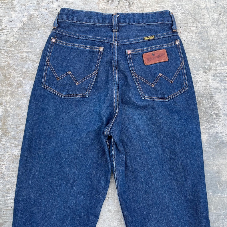K&ouml;nnte beinhalten: Dunkelblaue Jeans aus Denim mit dem Wrangler-Logo auf der Ges&auml;&szlig;tasche. Die Jeans haben eine hohe Taille, ein klassisches F&uuml;nf-Taschen-Design und kupferfarbene N&auml;hte. Die Ges&auml;&szlig;taschen haben die charakteristische 'W'-Naht von Wrangler.