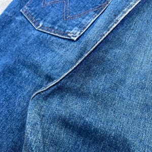 Pode incluir: Close-up de jeans azul denim, mostrando a textura e os detalhes da costura. A imagem destaca o bolso traseiro com um design de costura decorativa e as costuras da pe&ccedil;a. O tecido parece desgastado e desbotado.