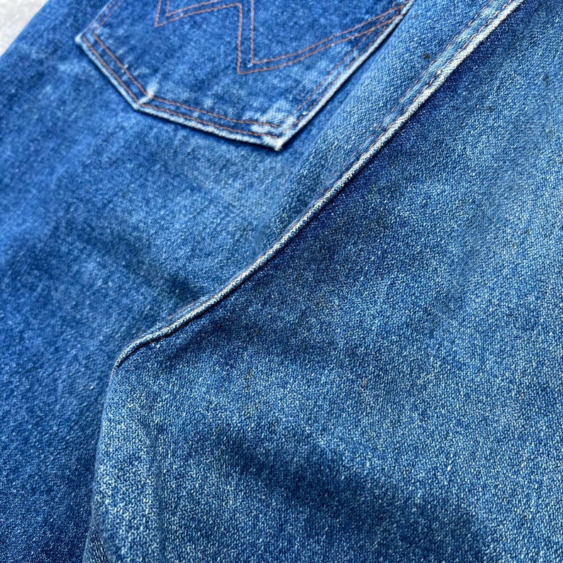 Pode incluir: Close-up de jeans azul denim, mostrando a textura e os detalhes da costura. A imagem destaca o bolso traseiro com um design de costura decorativa e as costuras da pe&ccedil;a. O tecido parece desgastado e desbotado.