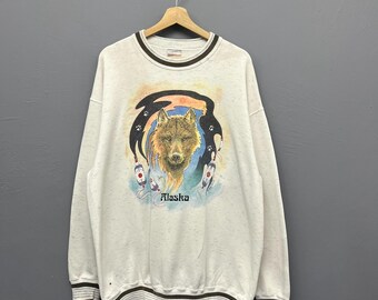 May include: Cream-colored sweatshirt with a wolf graphic and the word "Alaska". The wolf is depicted with detailed fur and surrounded by a black and orange design. The sweatshirt has brown stripes on the collar, cuffs, and hem.