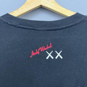 May include: A black crewneck sweatshirt featuring an embroidered red signature that reads Andy Warhol and a white embroidered double X logo positioned to the right. The garment is displayed on a light-coloured wooden hanger against a plain background.