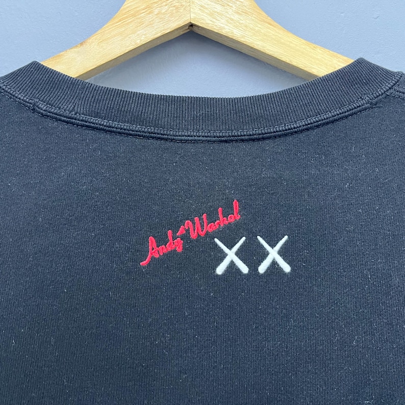 May include: A black crewneck sweatshirt featuring an embroidered red signature that reads Andy Warhol and a white embroidered double X logo positioned to the right. The garment is displayed on a light-coloured wooden hanger against a plain background.