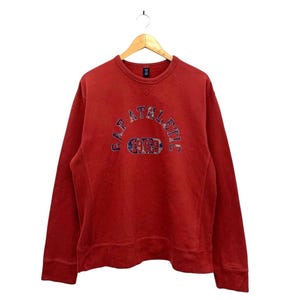 Może przedstawiać: Czerwona bluza Gap Athletic z długimi rękawami. Bluza ma napis "GAP ATHLETIC 1969" w wielokolorowym, łukowatym wzorze na piersi. Bluza wisi na drewnianym wieszaku na białym tle.