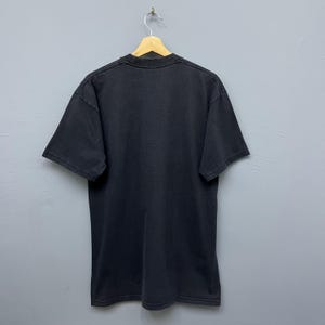 Op de afbeelding: Een zwart T-shirt met korte mouwen, hangend aan een houten hanger. Het T-shirt met ronde hals is gemaakt van een zacht materiaal. Het shirt wordt gepresenteerd tegen een grijze achtergrond.