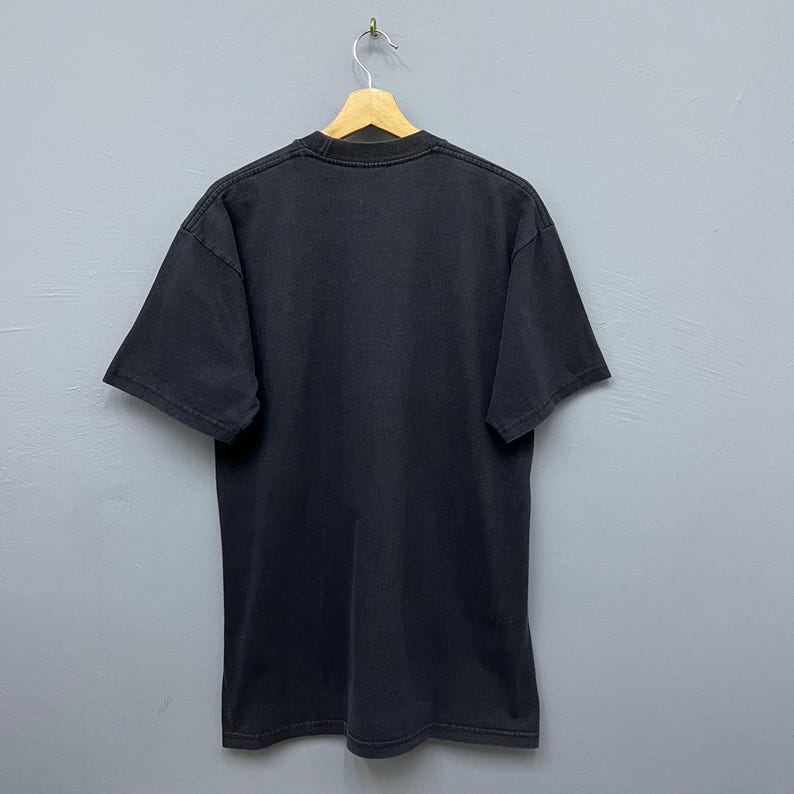 Op de afbeelding: Een zwart T-shirt met korte mouwen, hangend aan een houten hanger. Het T-shirt met ronde hals is gemaakt van een zacht materiaal. Het shirt wordt gepresenteerd tegen een grijze achtergrond.