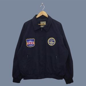 Puede incluir: Chaqueta azul marino con cuello y cierre de cremallera. La chaqueta presenta dos parches bordados: uno con estrellas y rayas y el texto "WE REMEMBER 1945-1975", y otro con un diseño circular.