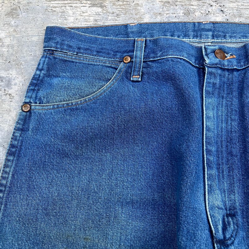 May include: Close-up of blue denim jeans. The jeans have a classic five-pocket design with copper-colored rivets and a button closure. The denim has a faded appearance, suggesting wear and washing. The jeans are a medium wash blue.
