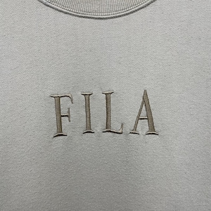 May include: A light gray crewneck sweatshirt featuring the word FILA embroidered in a dark gray thread across the center of the chest. The garment has a ribbed neckline and a smooth fabric texture, showcasing the brand name in a serif font.