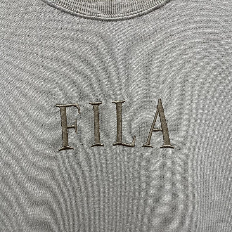 May include: A light gray crewneck sweatshirt featuring the word FILA embroidered in a dark gray thread across the center of the chest. The garment has a ribbed neckline and a smooth fabric texture, showcasing the brand name in a serif font.
