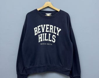 Puede incluir: Sudadera azul marino con la inscripción blanca "BEVERLY HILLS RODEO DRIVE". La sudadera de cuello redondo está colgada de una percha de madera sobre un fondo gris.