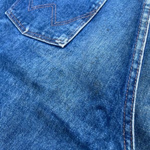 Pode incluir: Close-up de tecido denim azul. A imagem mostra um bolso com costuras laranja e uma parte de uma costura. O denim tem uma apar&ecirc;ncia desbotada com manchas mais escuras, sugerindo desgaste. A textura do tecido &eacute; vis&iacute;vel.