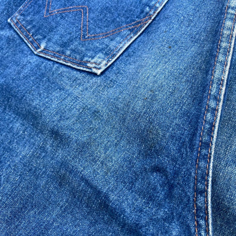 Pode incluir: Close-up de tecido denim azul. A imagem mostra um bolso com costuras laranja e uma parte de uma costura. O denim tem uma apar&ecirc;ncia desbotada com manchas mais escuras, sugerindo desgaste. A textura do tecido &eacute; vis&iacute;vel.