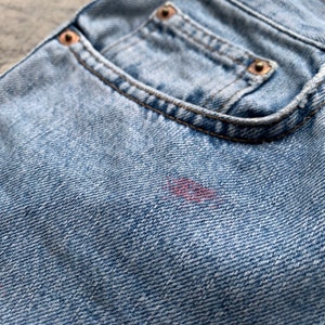 K&ouml;nnte beinhalten: Nahaufnahme einer hellblauen Jeans-Hosentasche mit Kupfernieten und einem kleinen Fleck. Die Tasche ist mit braunem Faden gen&auml;ht. Der Denim hat ein strukturiertes Aussehen.