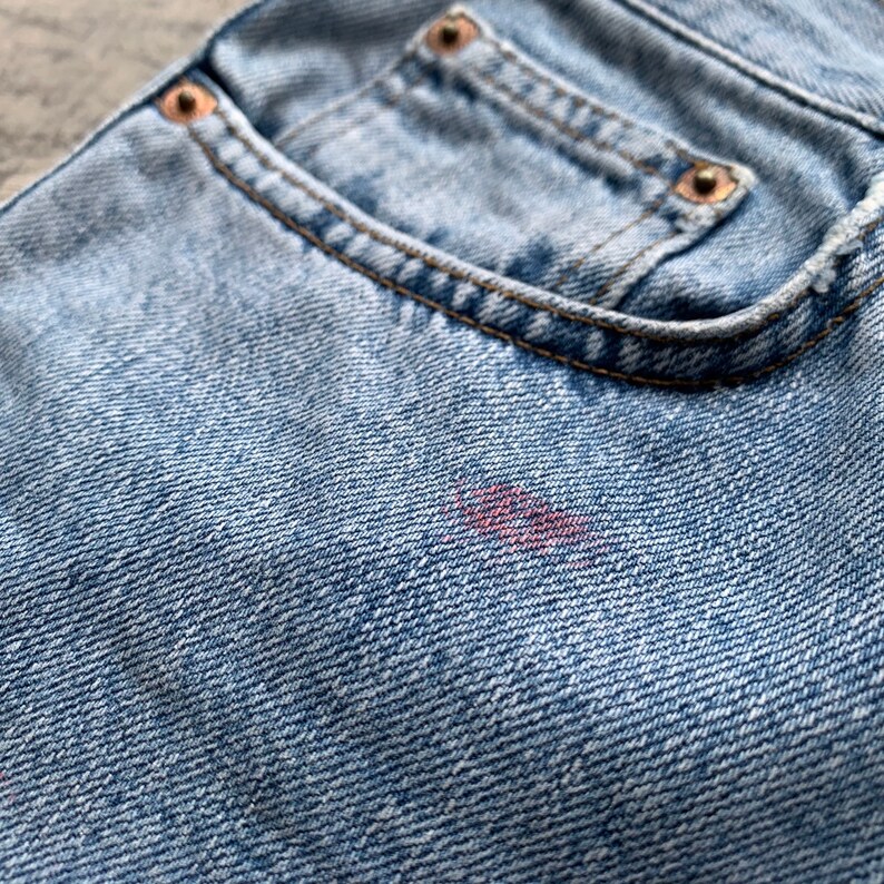 K&ouml;nnte beinhalten: Nahaufnahme einer hellblauen Jeans-Hosentasche mit Kupfernieten und einem kleinen Fleck. Die Tasche ist mit braunem Faden gen&auml;ht. Der Denim hat ein strukturiertes Aussehen.