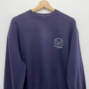 May include: A navy blue sweatshirt with a crew neck. The sweatshirt features a white graphic on the upper chest that reads "sport futur" above the letters "QC" and the word "courr&egrave;ges paris" below. The sweatshirt is hanging on a wooden hanger.