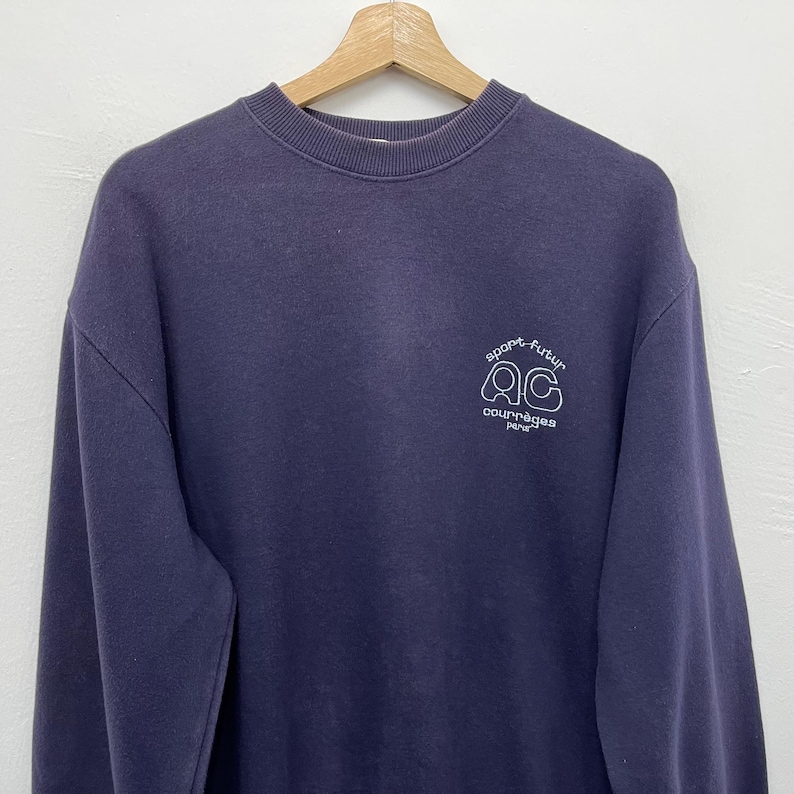 May include: A navy blue sweatshirt with a crew neck. The sweatshirt features a white graphic on the upper chest that reads "sport futur" above the letters "QC" and the word "courr&egrave;ges paris" below. The sweatshirt is hanging on a wooden hanger.