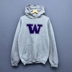 May include: A light gray hooded sweatshirt featuring a large, bold purple letter W with a white outline centered on the chest. The pullover garment includes a front kangaroo pocket, long sleeves with ribbed cuffs, and a matching ribbed hemline.