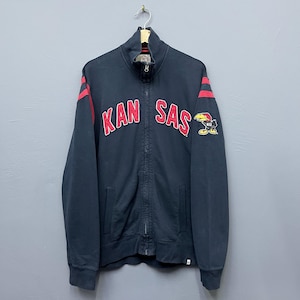 May include: A navy blue zip-up jacket with red stripes on the sleeves and the text "KAN SAS" in red. A patch with a bird logo is on the right sleeve. The jacket has a high collar and two front pockets.