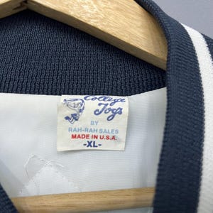 May include: Close-up of a vintage navy blue and white varsity jacket. The jacket features a white lining and a label that reads "College Jogs by Rah-Rah Sales Made in U.S.A. -XL-". The jacket is hanging on a wooden hanger.