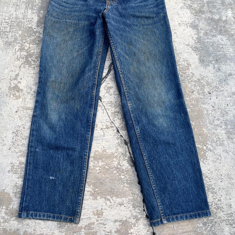 Pode incluir: Cal&ccedil;as de ganga azuis com um corte reto cl&aacute;ssico. As cal&ccedil;as de ganga mostram sinais de desgaste, com &aacute;reas desbotadas e desgaste subtil. A cor geral &eacute; azul m&eacute;dio, com um design tradicional de cinco bolsos e um fecho de bot&atilde;o e fecho de correr.