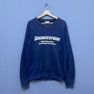 以下が含まれることがあります： 白い文字で「BRIDGESTONE Performance Gear for the Super Contenders」と書かれたネイビーブルーのスウェットシャツ。スウェットシャツは木製のハンガーに吊るされており、青い背景にあります。クルーネックと長袖です。