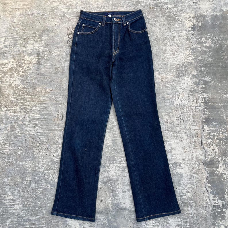May include: Dark wash denim jeans with a high waist and straight leg. The jeans have classic five-pocket styling with yellow stitching and a button and zipper closure. The jeans are a solid dark blue color.